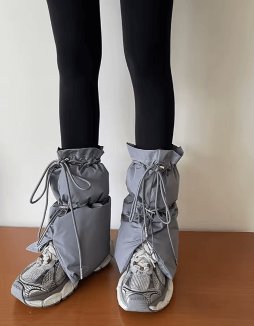 A person wearing black leggings and gray quilted puffy boot covers over sneakers, tied with adjustable laces, standing indoors against a plain wall.