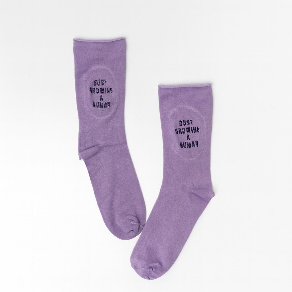 Flat lay of a pair of lilac maternity socks with black embroidery text that reads “Busy Growing a Human.