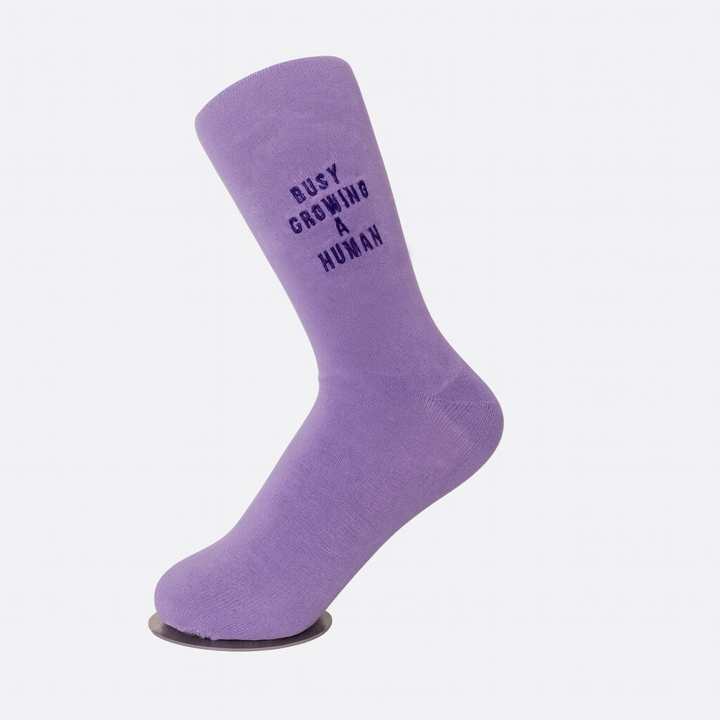 Single lilac sock displayed on a mannequin foot, showing black embroidered text “Busy Growing a Human.