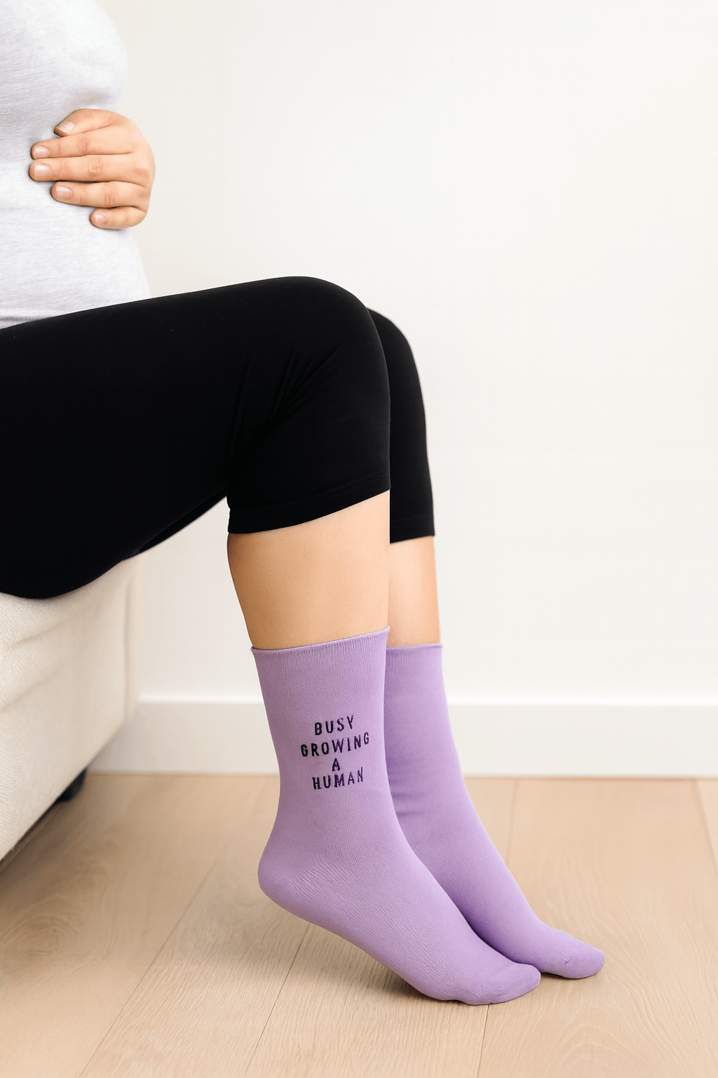 Pregnant woman sitting on a couch, wearing black leggings and lilac socks embroidered with the words “Busy Growing a Human.
