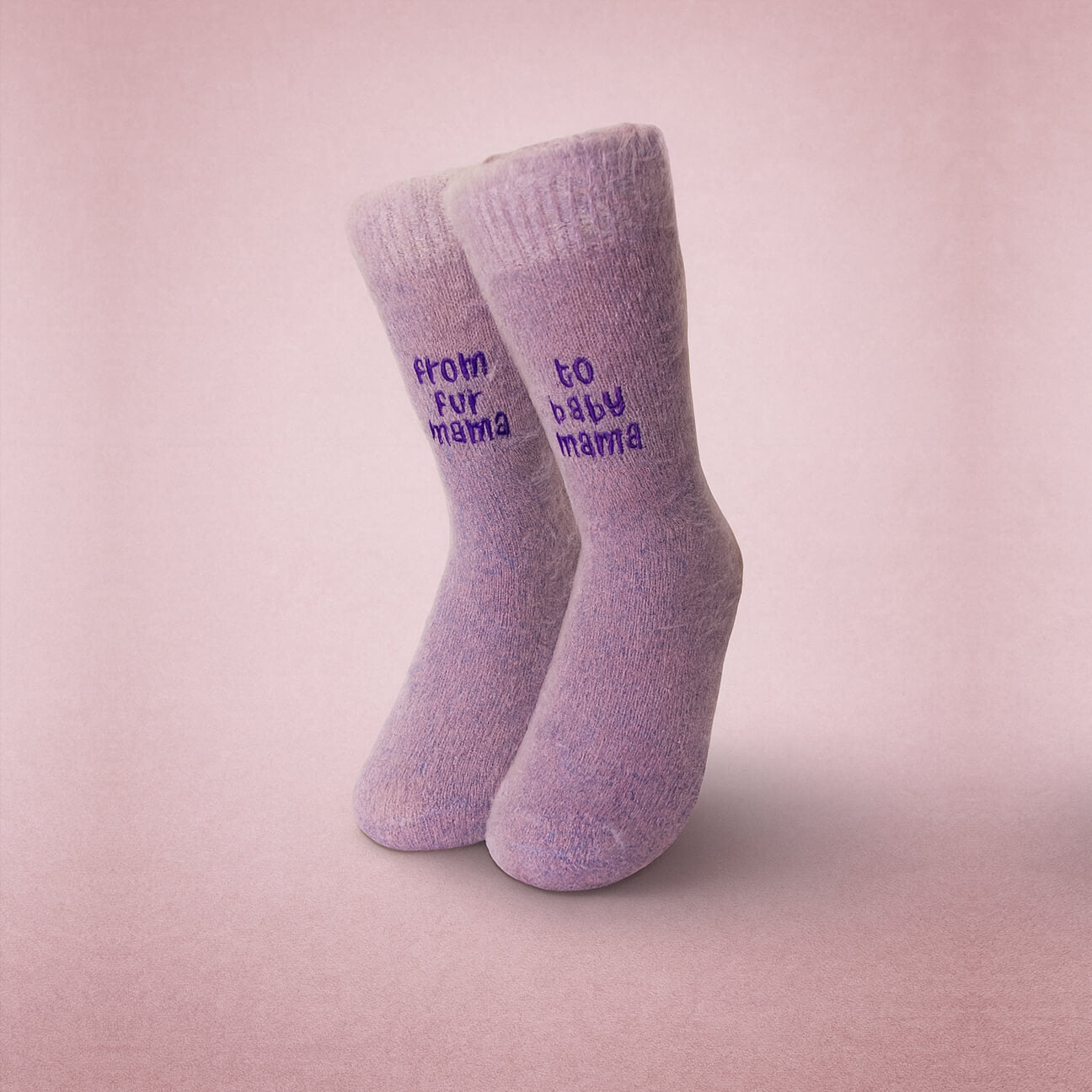 Pair of fuzzy lilac socks standing upright on a soft pink background. One sock reads “from fur mama” and the other reads “to baby mama” in purple embroidery