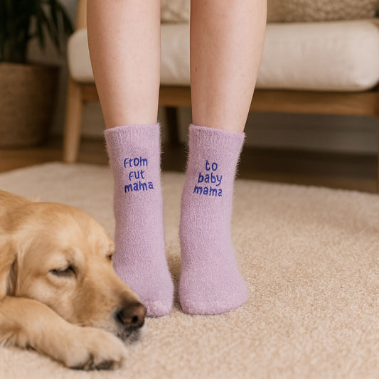 Woman wearing fuzzy lilac socks embroidered with “from fur mama” and “to baby mama,” standing on a beige carpet beside a sleeping golden retriever dog.