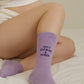 Pregnant woman sitting on a bed wearing lilac socks with the phrase “Busy Growing a Human” embroidered on the side.