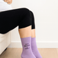 Pregnant woman sitting on a couch, wearing black leggings and lilac socks embroidered with the words “Busy Growing a Human.