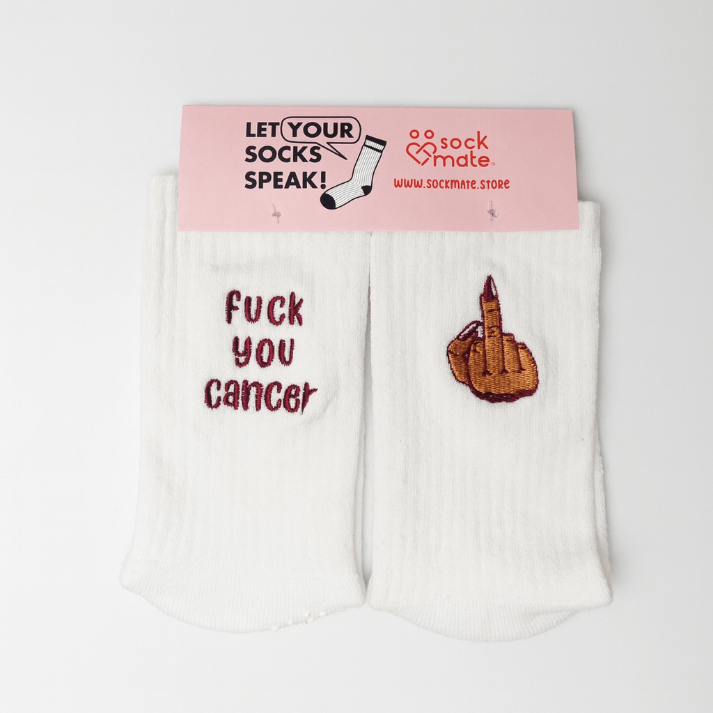 A pair of white embroidered socks from Sockmate, shown with pink branded packaging labeled “Let Your Socks Speak!” and “www.sockmate.store.” One sock reads “fuck you cancer” in red embroidery, and the other shows a hand with red nail polish giving the middle finger. Supportive and empowering gift socks for cancer fighters and survivors.