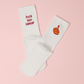 A pair of white non-slip socks displayed on a soft pink background. One sock is embroidered with the bold red phrase “fuck you cancer,” and the other features an embroidered hand with red nail polish giving the middle finger. Empowering and cheeky anti-cancer message socks designed for hospital stays or recovery comfort.