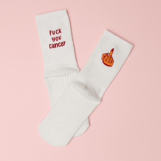 A pair of white non-slip socks displayed on a soft pink background. One sock is embroidered with the bold red phrase “fuck you cancer,” and the other features an embroidered hand with red nail polish giving the middle finger. Empowering and cheeky anti-cancer message socks designed for hospital stays or recovery comfort.