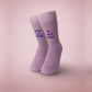 Pair of fuzzy lilac socks standing upright on a soft pink background. One sock reads “from fur mama” and the other reads “to baby mama” in purple embroidery