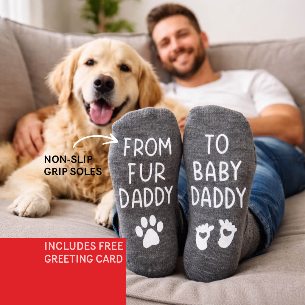 Man sitting on a couch with a dog, wearing socks that read 'From Fur Daddy to Baby Daddy' with non-slip grip soles.
