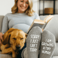 Woman sitting on a couch with a dog, wearing socks with humorous text "sorry I just can't today, i am growing a tiny human", in a cozy living room setting.